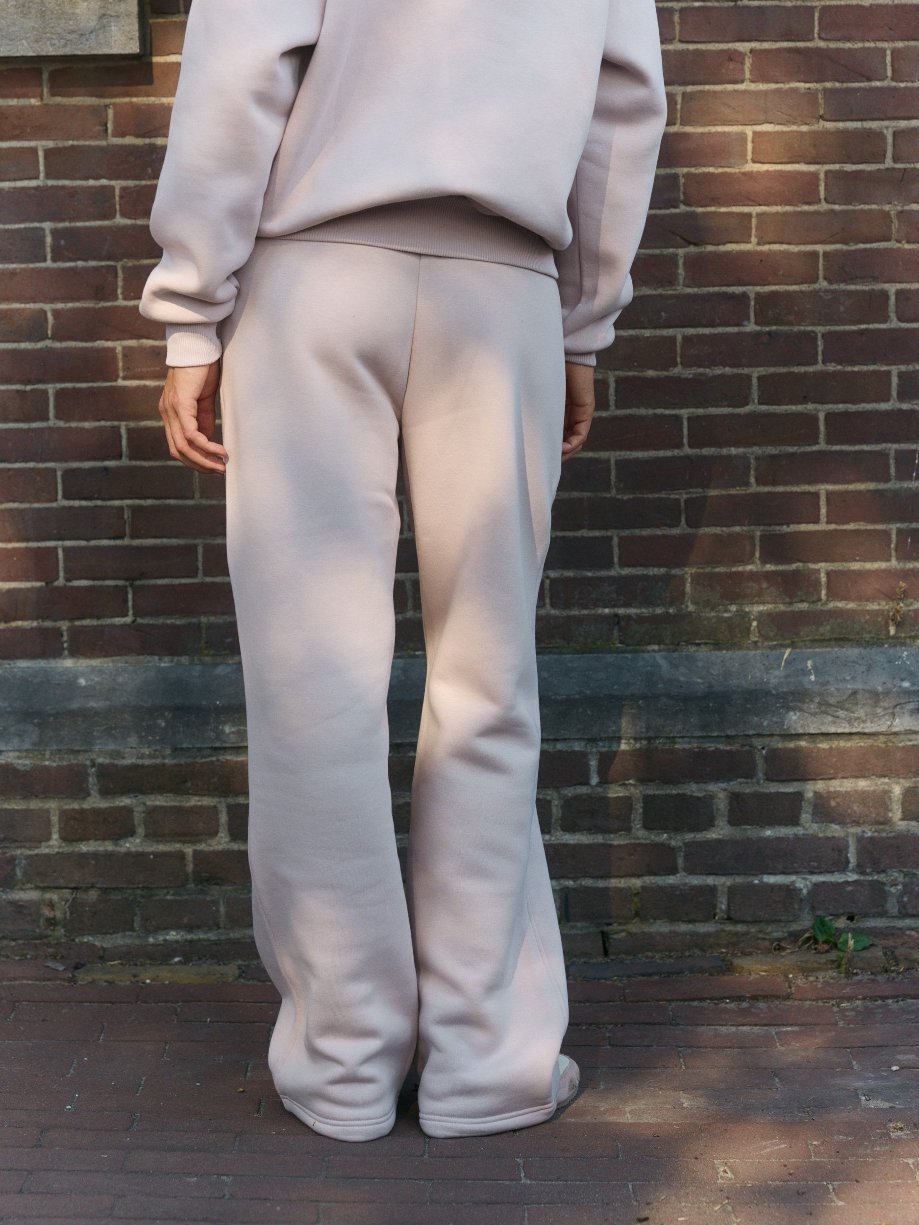 Back view of a woman wearing dusty pink straight-leg sweatpants against a brick wall.